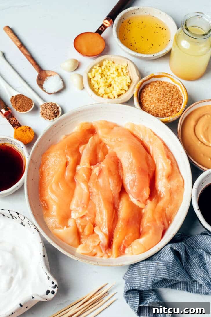 Pieces of raw chicken tender resting in a white mixing bowl, surrounded by various small bowls holding individual marinade ingredients like coconut milk, brown sugar, and spices.