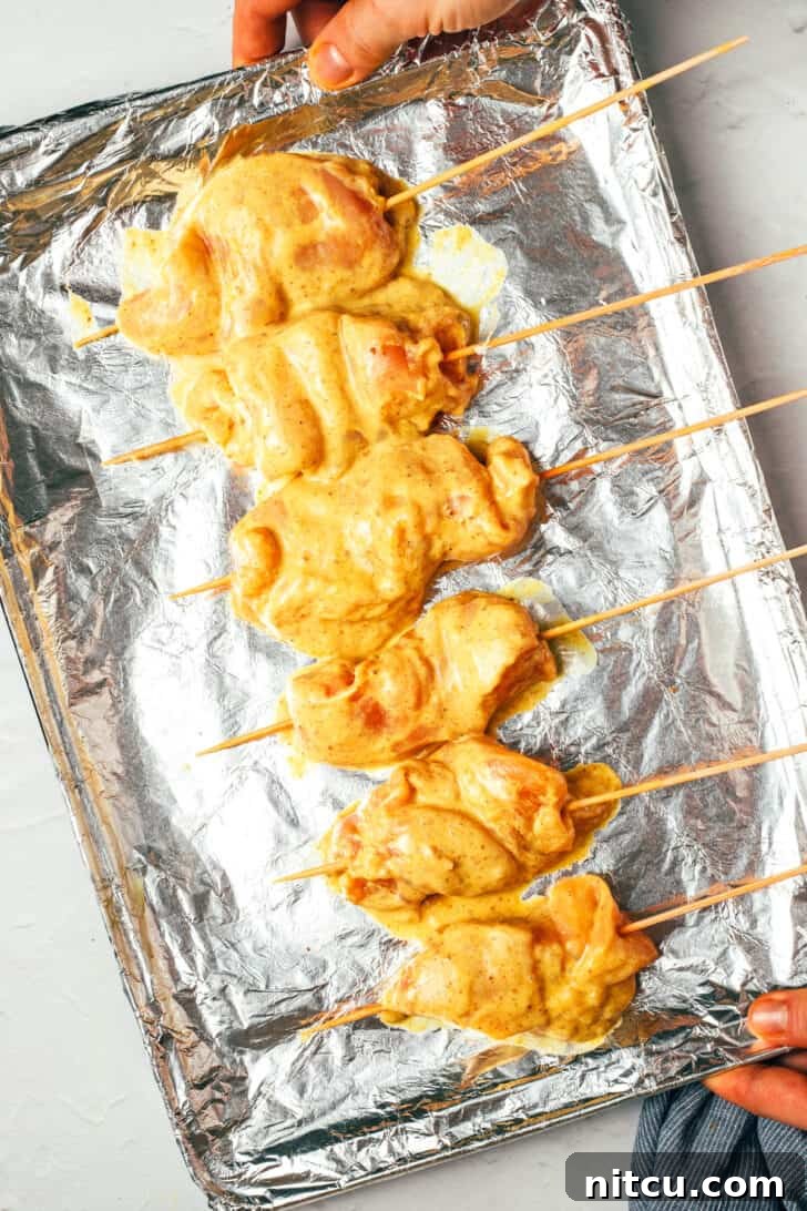 A hand holding a baking sheet covered with aluminum foil, showcasing six raw, perfectly threaded chicken skewers, ready for grilling.