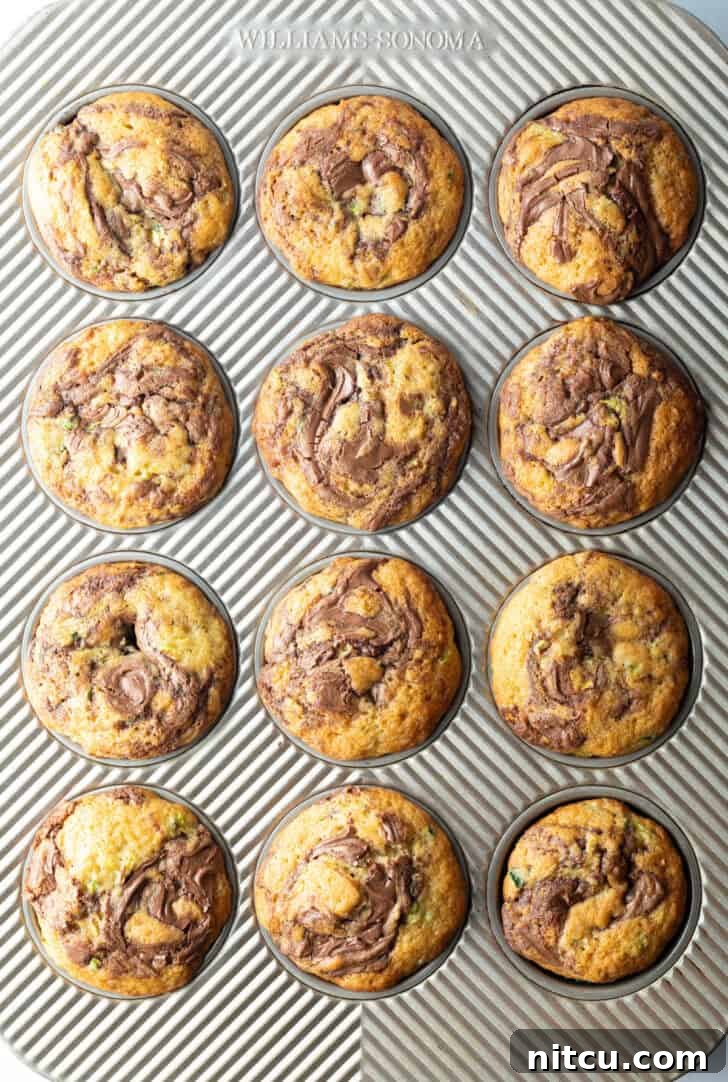 An inviting top-down view of a dozen perfectly golden-brown zucchini and Nutella muffins, still in their baking tin, showcasing their beautiful swirls and ready to be served.