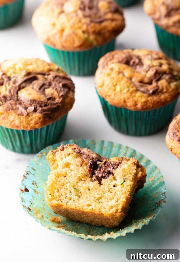 A vibrant display of a dozen freshly baked chocolate zucchini muffins, each adorned with a distinct Nutella swirl, perfectly arranged in green cupcake wrappers. One muffin is broken open to reveal its moist, tender interior and the enticing marbling of the chocolate and hazelnut.