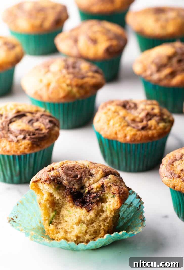 A close-up view of a dozen chocolate zucchini muffins with prominent Nutella swirls, presented in green cupcake wrappers. One muffin is thoughtfully split in half, revealing its moist interior and the beautiful marbling of the chocolate hazelnut spread.