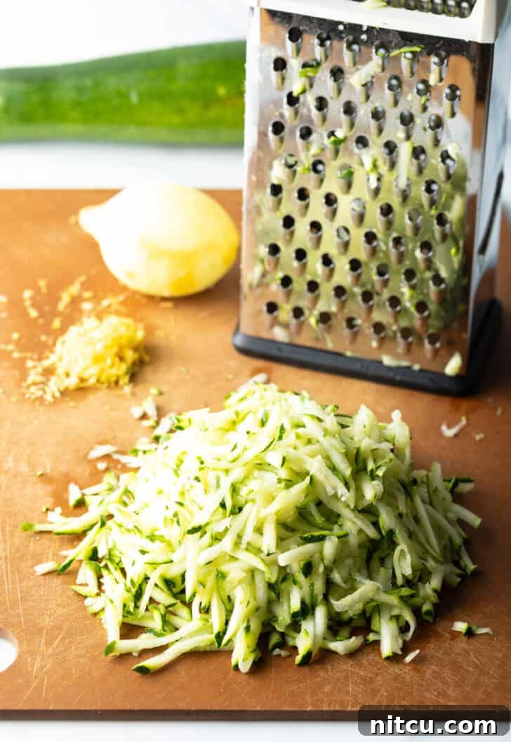 A box grater, a whole fresh lemon, and a generous pile of shredded zucchini neatly arranged on a rustic wood cutting board, showcasing the fresh ingredients.