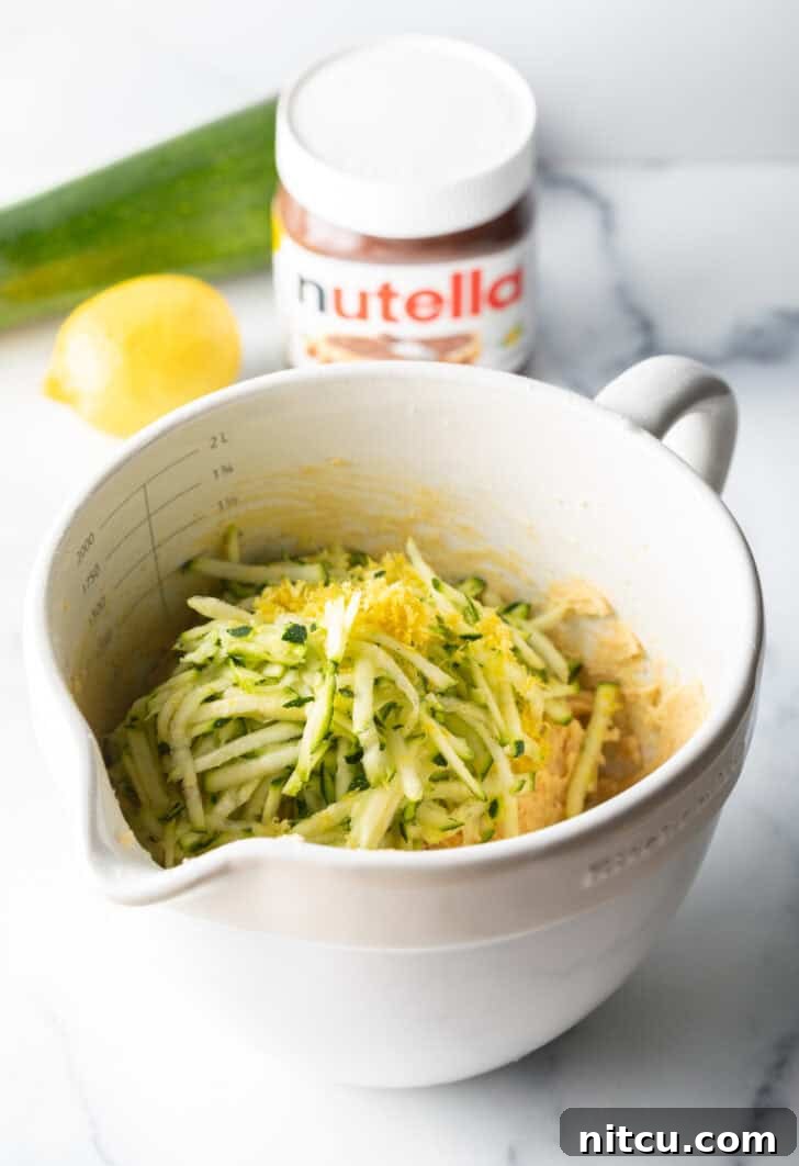 A white measuring bowl containing freshly shredded zucchini, brightened by the addition of aromatic lemon zest, ready for the muffin batter.