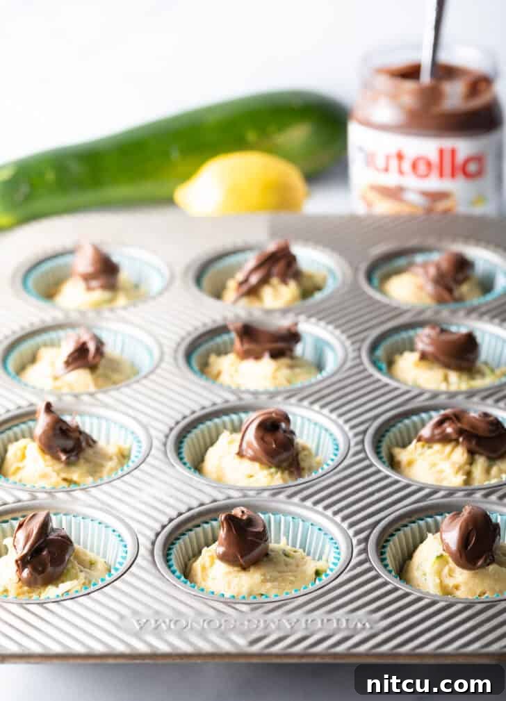 Unbaked zucchini muffin batter in a cupcake tin, each portion topped with a generous dollop of rich Nutella, poised for swirling.