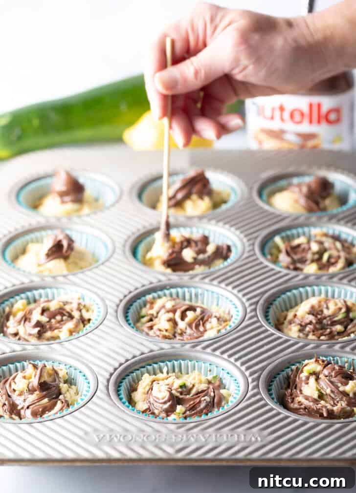 A hand skillfully using a wooden skewer to gently swirl Nutella into the unbaked zucchini muffin batter within a baking tin, creating beautiful marble patterns.
