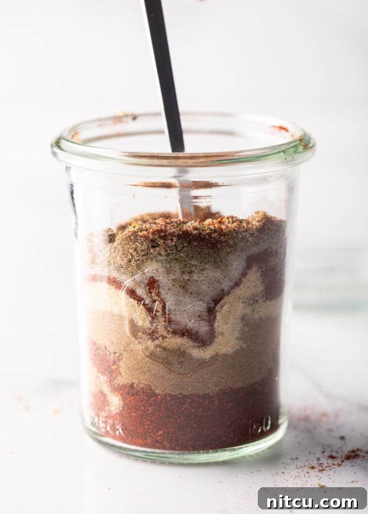 A spoon actively stirring layers of chili powder, cumin, garlic powder, and other spices in a glass jar, demonstrating the mixing process.