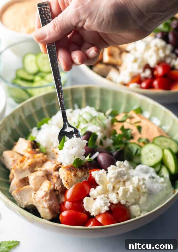 Fork grabbing a bite from a Greek chicken bowl with pieces of chicken, rice, sliced cherry tomatoes, kalamata olives, tzatziki sauce, cucumbers and feta cheese.