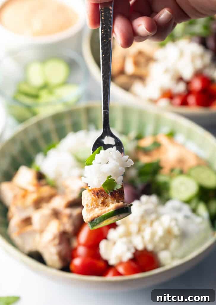 Greek chicken bowl with pieces of chicken, rice, sliced cherry tomatoes, kalamata olives, tzatziki sauce, cucumbers and feta cheese. Fork holding a bite of rice and cucumber to camera