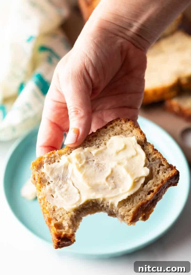 A hand holding a moist slice of banana bread, generously slathered with butter, showing a bite taken from the corner.