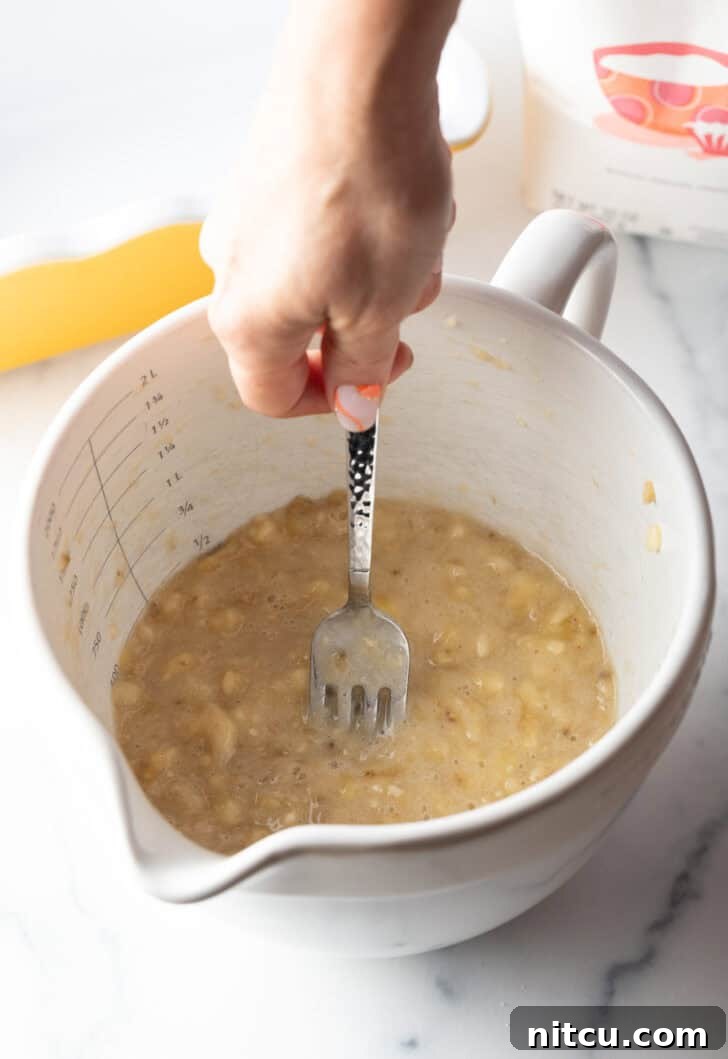 A hand using a metal fork to mash ripe bananas in a white mixing bowl, illustrating the mashing process.
