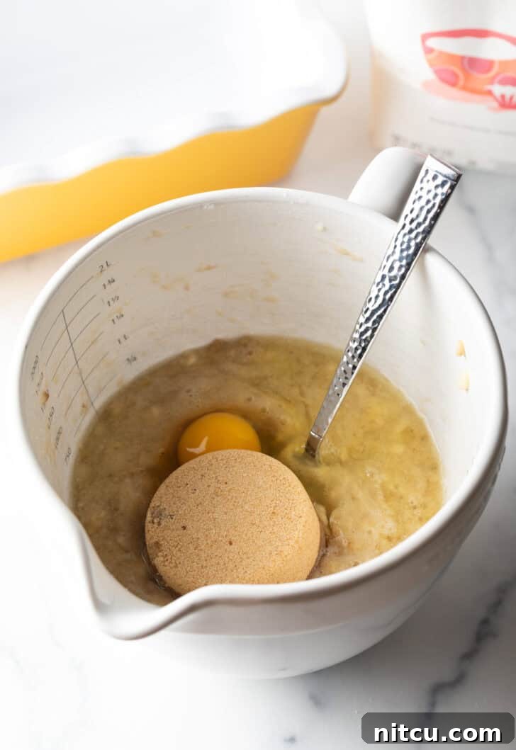 Adding a scoop of brown sugar and a raw egg into a large white mixing bowl already containing mashed bananas, showing the next step in the recipe.