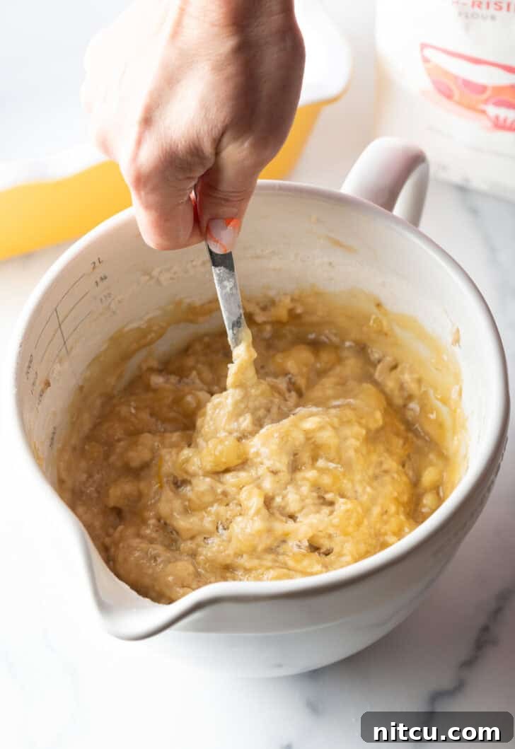 A hand using a metal spoon to gently stir the banana bread batter in a large white mixing bowl, demonstrating the mixing technique.
