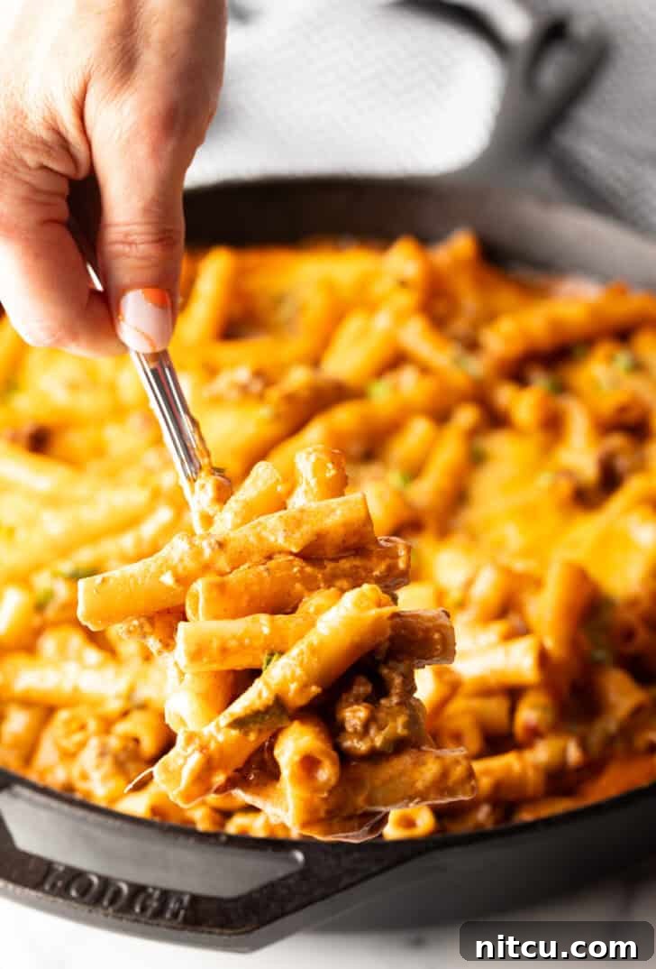 Hand using a large spoon to scoop noodle cheeseburger casserole from a large skillet towards camera. The casserole is steaming and freshly cooked.