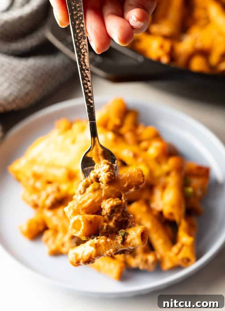 Hand holding a forkful of hamburger casserole to camera. The cheesy noodles and beef are prominent.