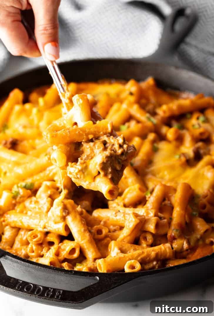 Hand using a large spoon to scoop noodle cheeseburger casserole from a large skillet towards camera. The rich, cheesy, and saucy texture is visible.