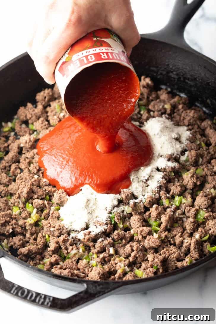 Hand adding canned tomato sauce to a skillet of cooked ground beef and ranch seasoning mix. The sauce is rich red, contrasting with the browned meat.