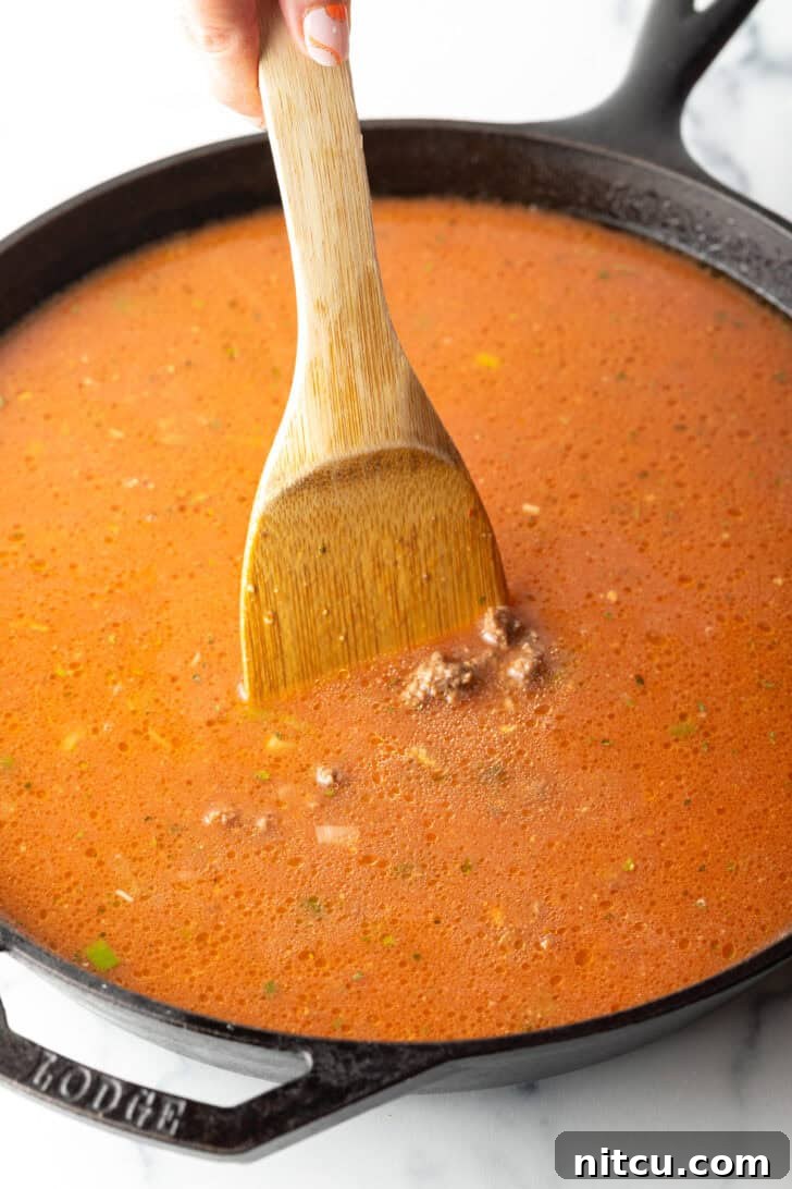 Using a wood spatula to stir tomato and beef mixture in a cast iron skillet. The mixture is simmering gently.
