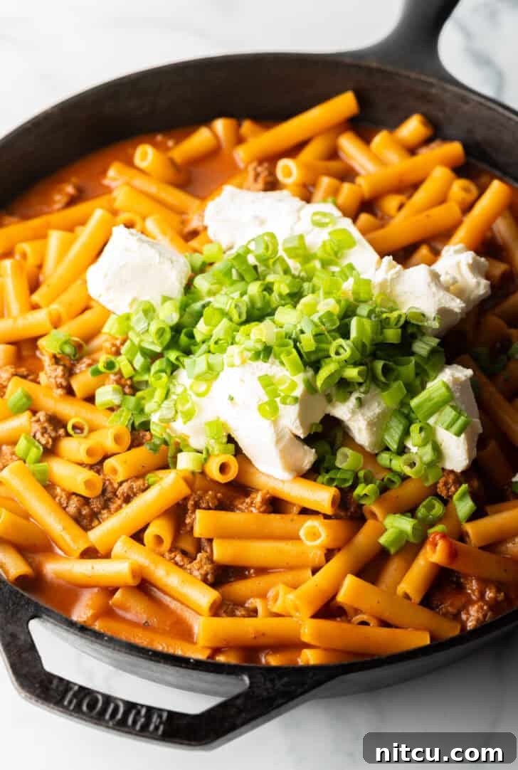 Adding cream cheese and chopped green onions to a cast iron skillet with ground beef and noodles. The cream cheese is cubed for quick melting.