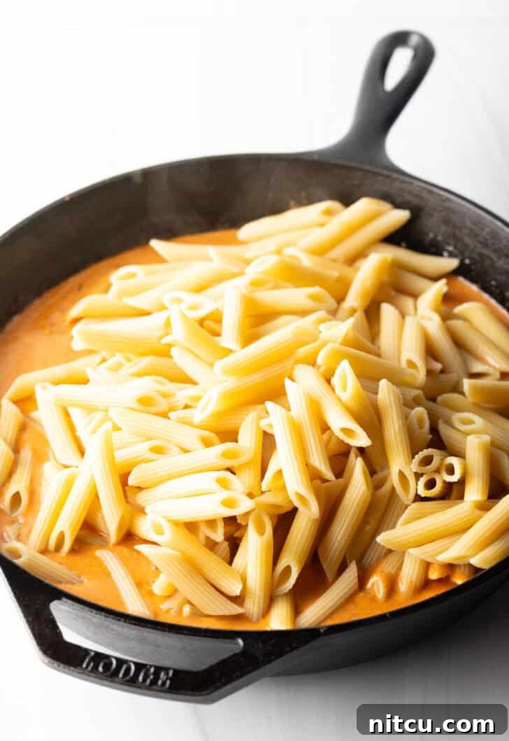 Adding penne noodles to the velvety cream sauce in a cast iron skillet.