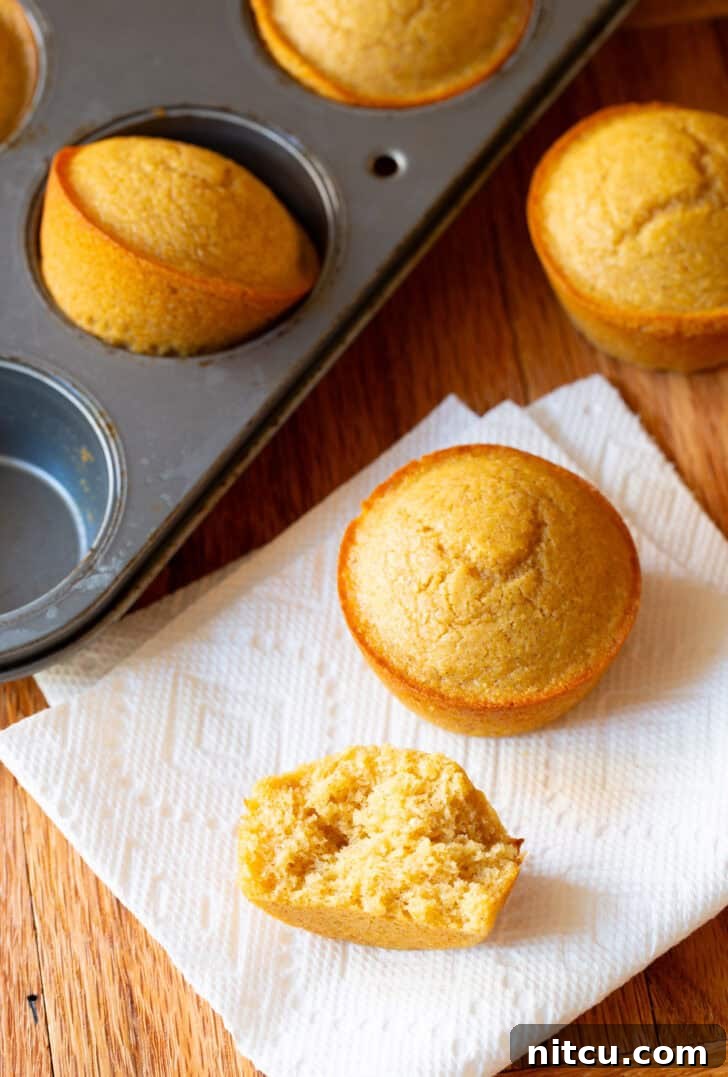 Two golden-brown honey cornbread muffins on a white napkin, one expertly split in half to showcase its incredibly moist, fluffy interior, with a full muffin tin in the background.