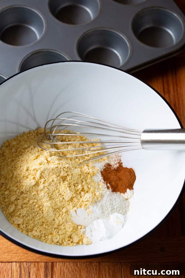 Dry ingredients for honey cornbread muffins, including cornmeal, flour, and cinnamon, perfectly blended in a white mixing bowl with a whisk.