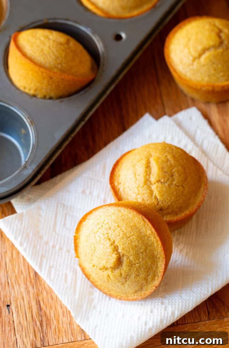 Two warm honey cornbread muffins on a white napkin, nestled beside a muffin baking tin overflowing with more freshly baked, golden muffins.