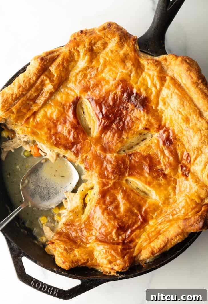 A golden brown puff pastry topped chicken pot pie in a cast iron skillet. A serving has been removed, revealing the creamy filling.