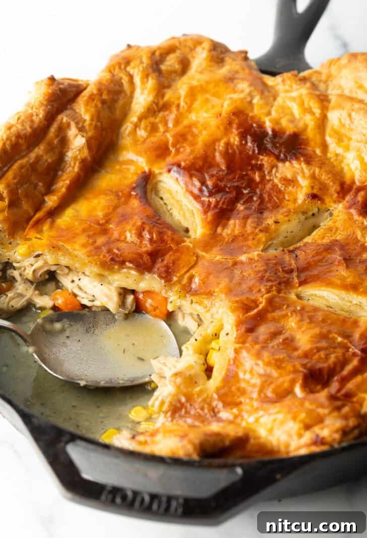 A cast iron skillet of chicken pot pie topped with golden brown puff pastry, with a large serving spoon having removed a quarter portion.