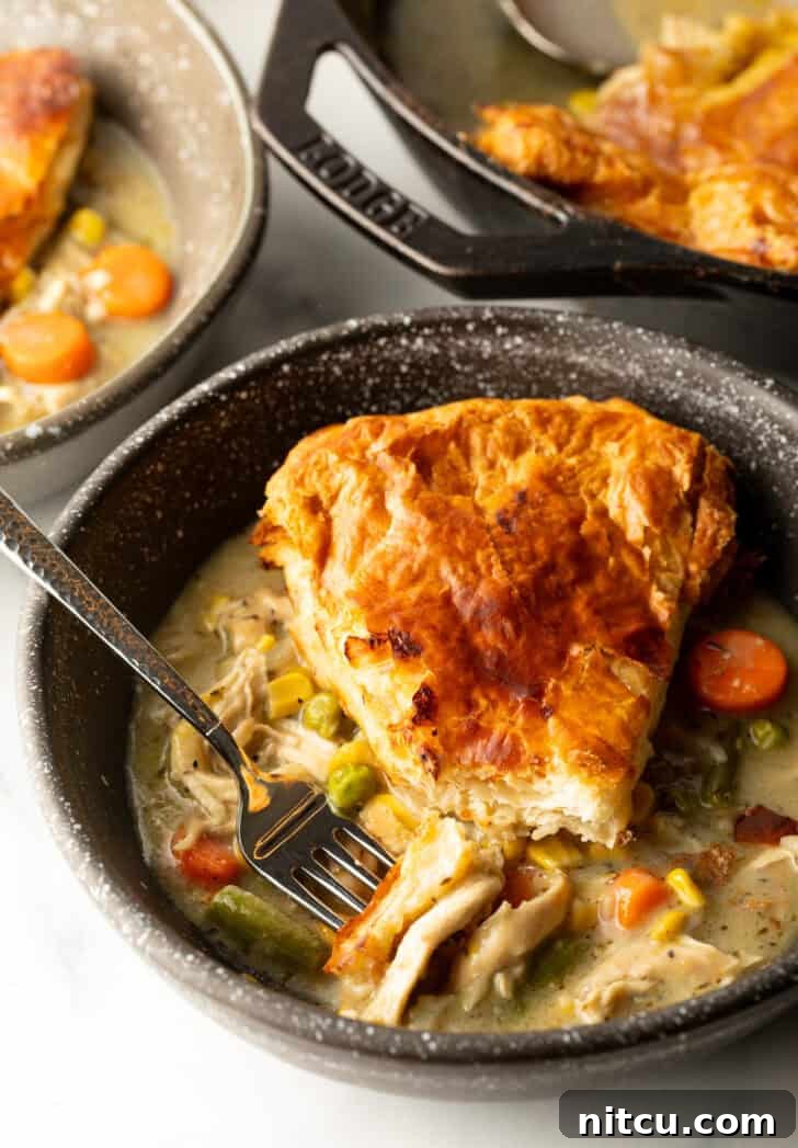 Top down view of a wedge of chicken pot pie with puff pastry crust on a black plate with a fork, ready to be eaten.