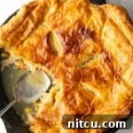 Skillet of chicken pot pie topped with golden brown puff pastry. A serving spoon has removed a quarter portion.
