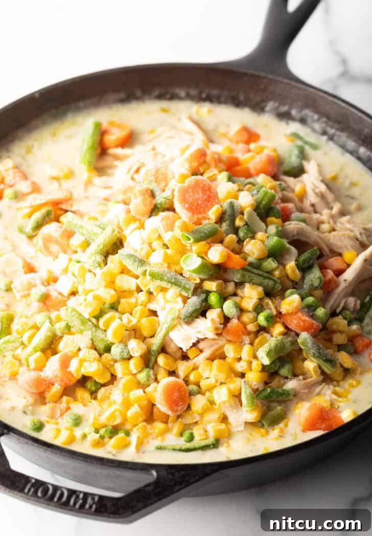 Adding frozen mixed vegetables to a rich, creamy gravy in a cast iron skillet.