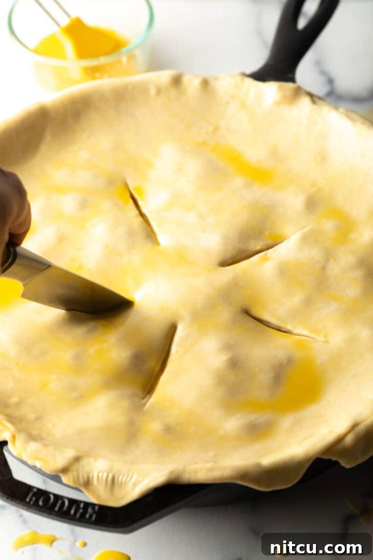 A chef's knife cutting steam vents into the uncooked puff pastry crust atop a pie.