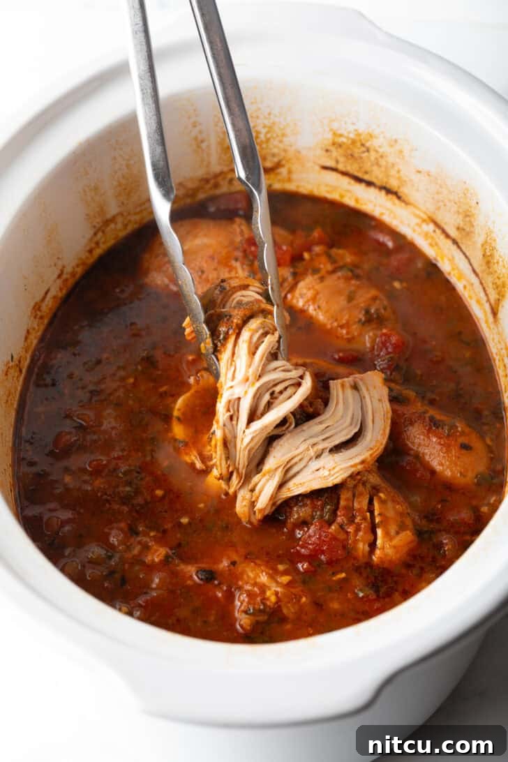 Tongs holding up shredded chicken above a white slow cooker.
