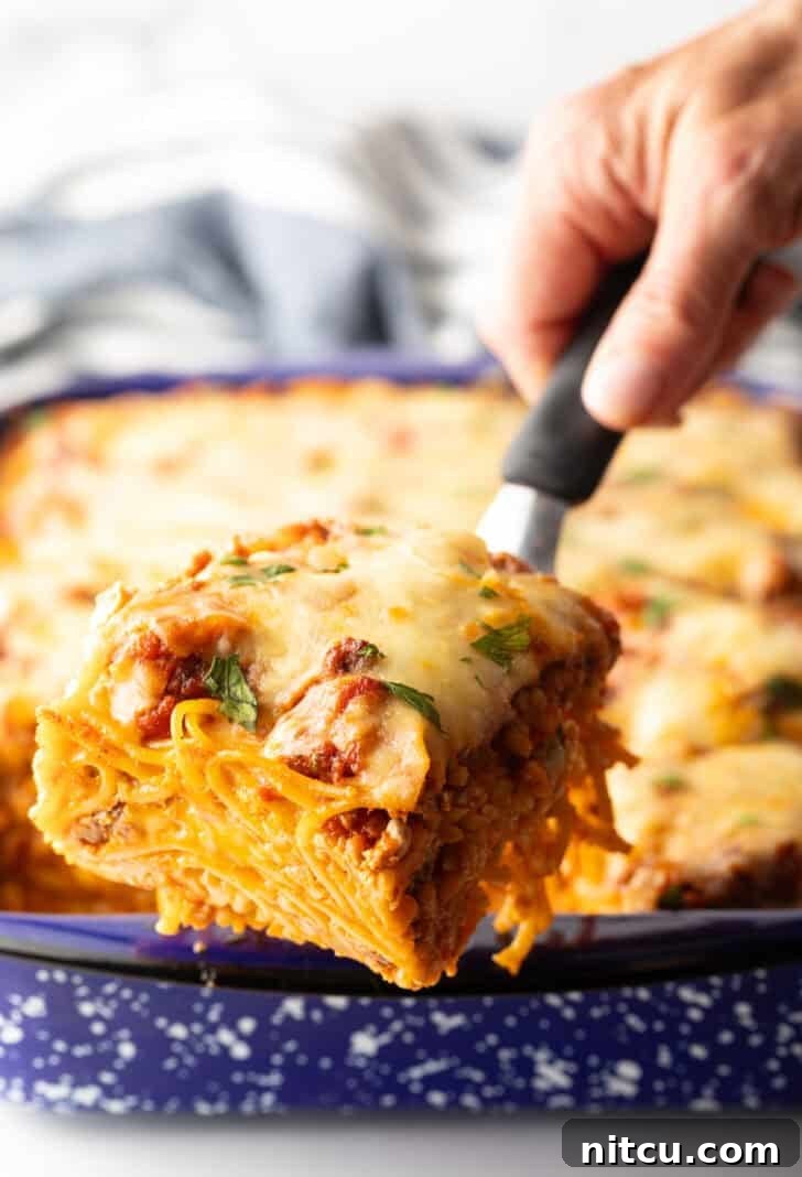 Hand with a spatula holding a serving of baked spaghetti to camera, showcasing melted cheese and rich sauce.
