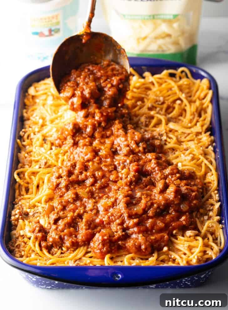 A ladle pouring the remaining meat sauce over the spaghetti base in a blue casserole dish.