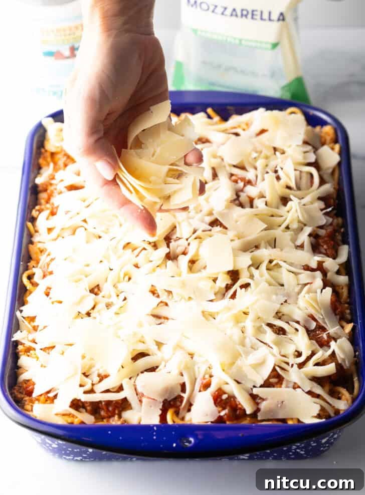 Hand adding shredded parmesan cheese on top of the spaghetti casserole, completing the cheesy layer.