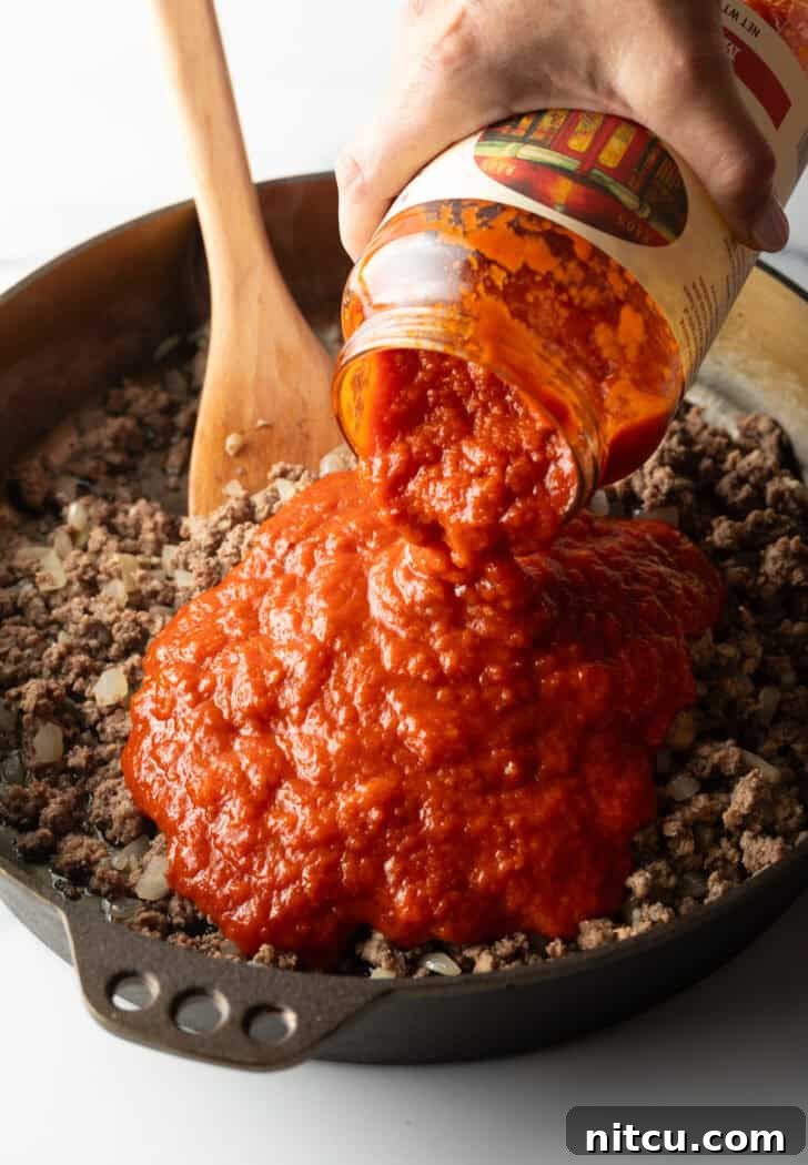 Adding jarred marinara sauce to a skillet with cooked ground meat and aromatics.