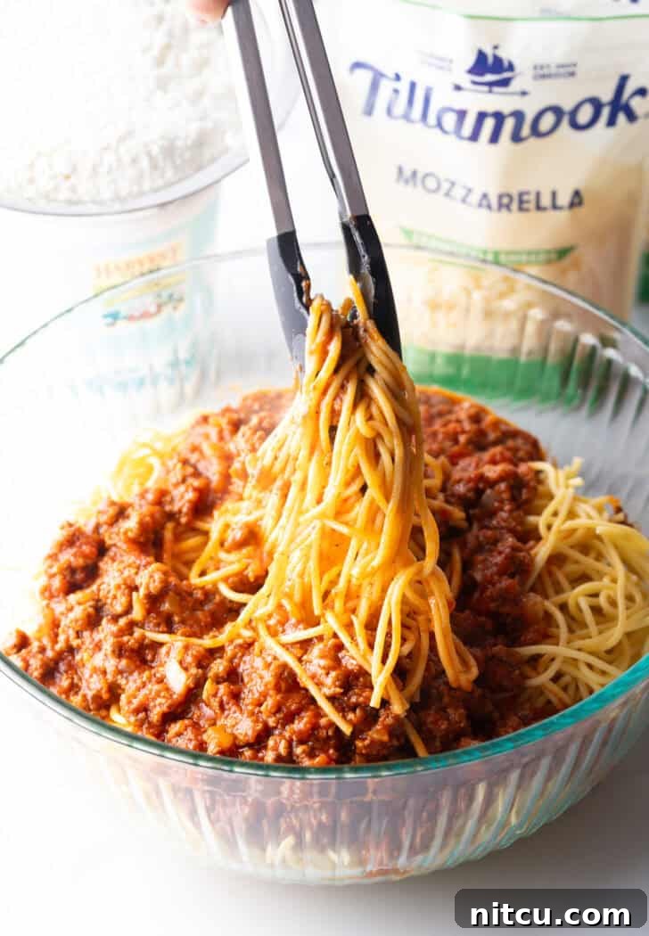 Tongs tossing cooked spaghetti together with half of the meat sauce in a large glass bowl.