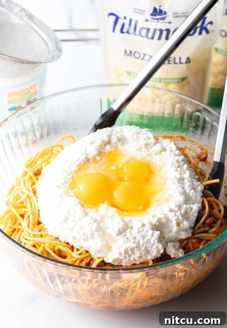 Glass bowl with cooked spaghetti, cottage cheese, and three cracked eggs in the middle, ready to be mixed.
