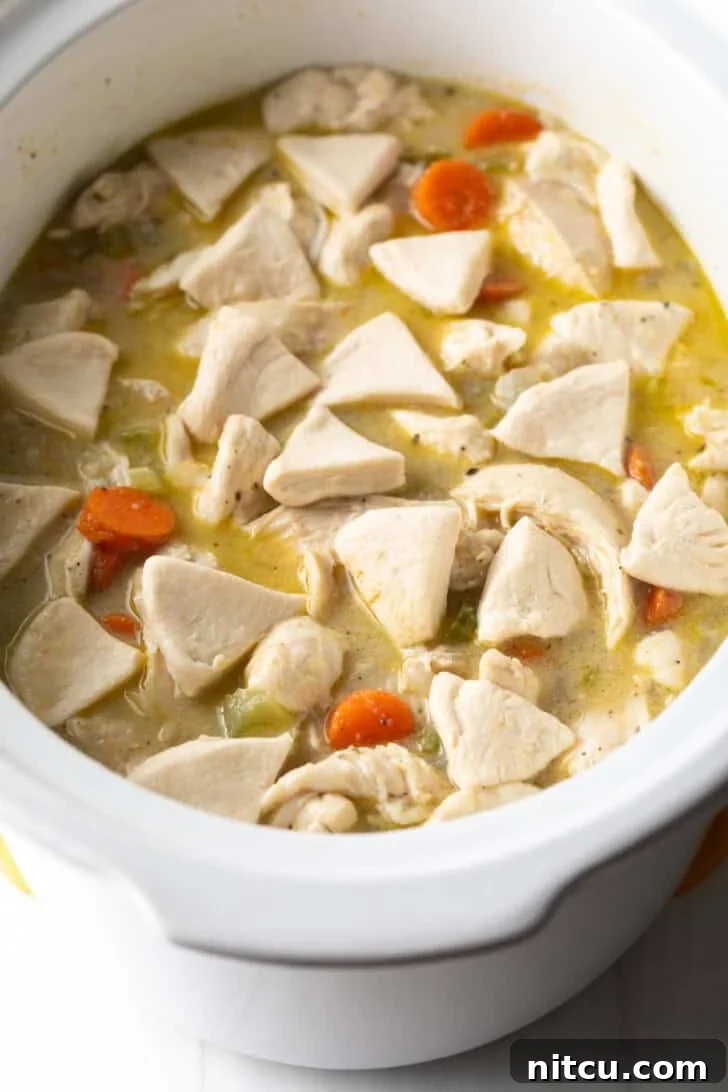 Triangular biscuit dough pieces gently dropped into a white slow cooker containing shredded chicken and a rich, simmering broth.