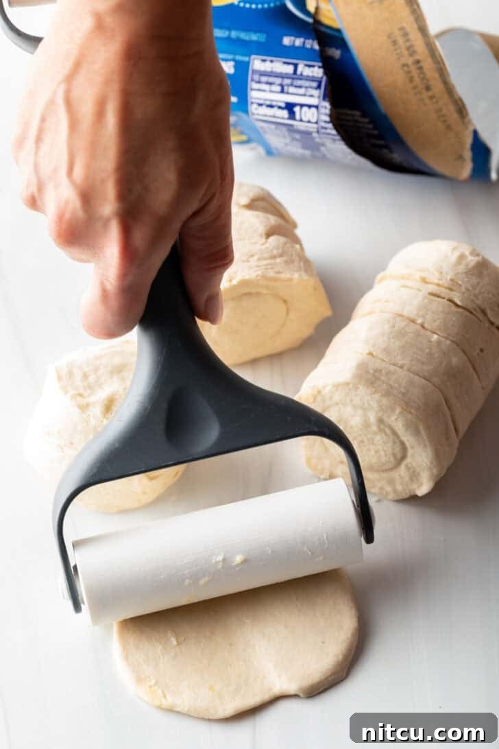 A hand using a rolling pin to press a raw biscuit flat on a cutting board, preparing it for slicing into dumplings.