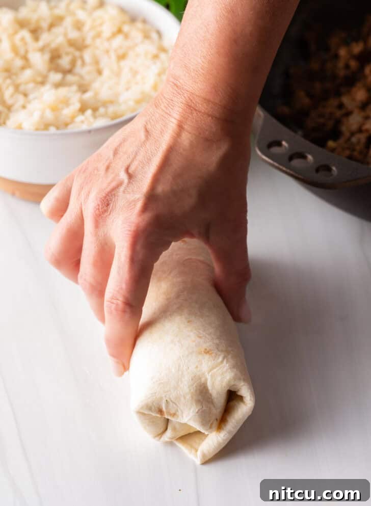 Hand wrapping a beef burrito, showing the almost completed, tight roll.