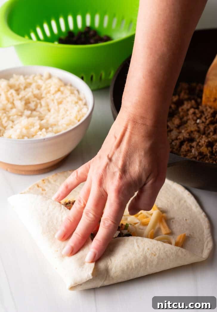 Hand folding a tortilla to make a burrito filled with beef and cheese, demonstrating the initial fold.