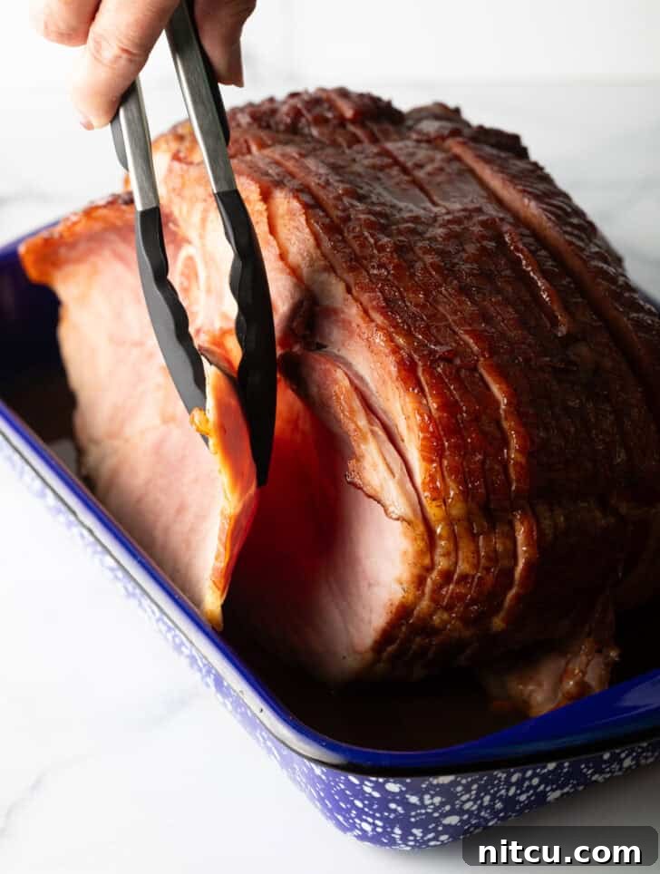 Tongs gently removing the front slice of a perfectly cooked spiral ham from a baking dish, showcasing its tenderness.