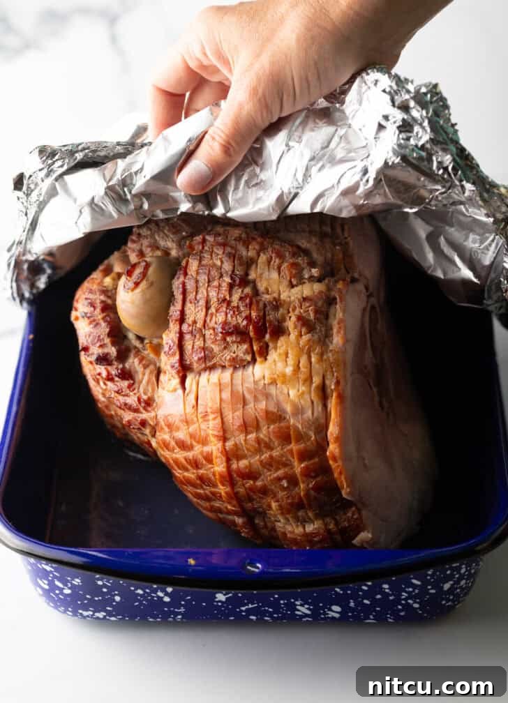 A cook carefully covering a spiral ham in a blue and white speckled roasting pan with aluminum foil, illustrating the step to lock in moisture.