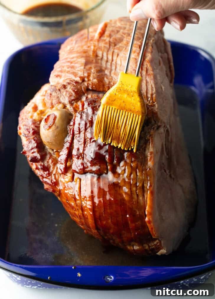 A cooked spiral ham being basted with a rich, golden sauce using a yellow silicone brush, highlighting the glazing process.