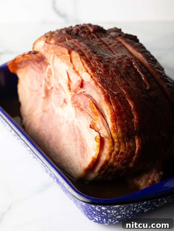 A cooked spiral ham, glistening with glaze, in a blue and white speckled roasting pan, with a few slices already carved.