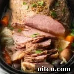 Sliced whole corned beef surrounded by large pieces of carrots, cabbage, and baby potatoes in a black crock pot.