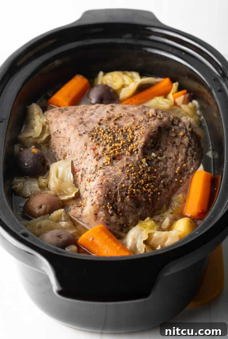 Whole corned beef surrounded by large pieces of carrots, cabbage, and baby potatoes in a black crock pot.
