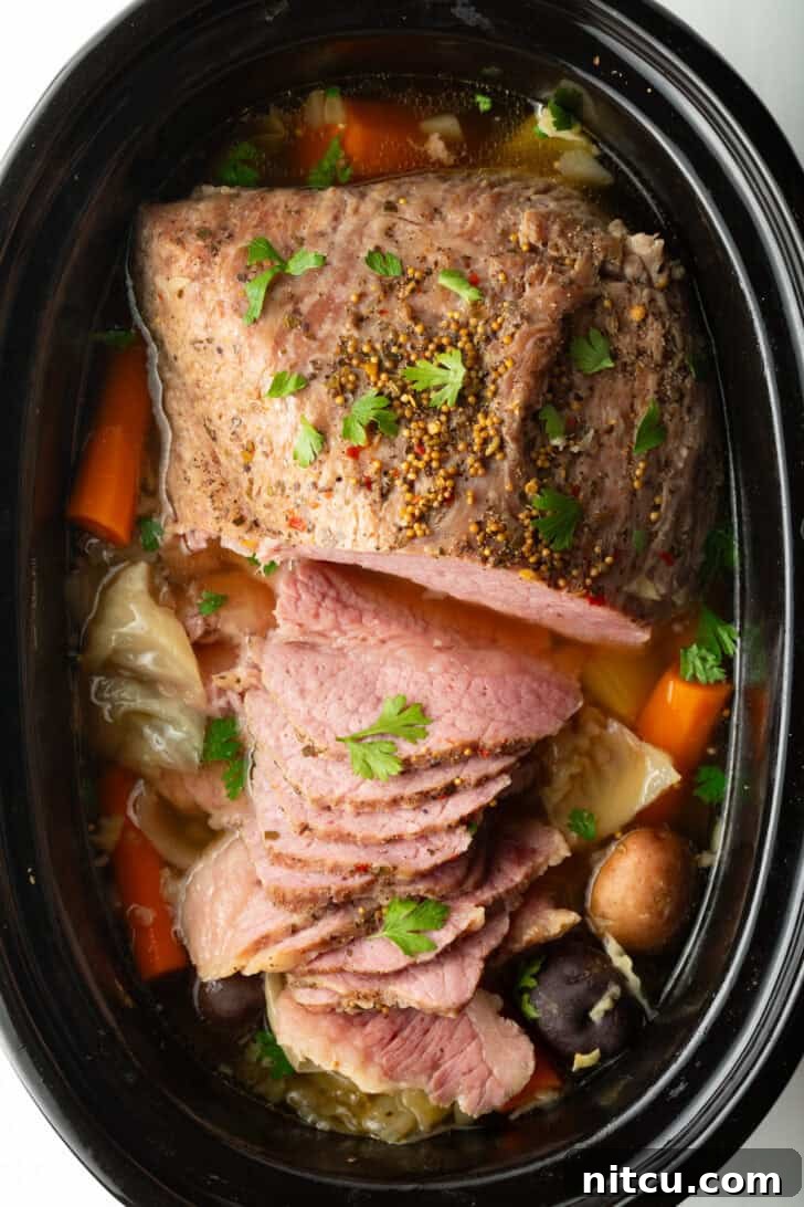 Sliced whole corned beef surrounded by large pieces of carrots, cabbage, and baby potatoes in a black crock pot.